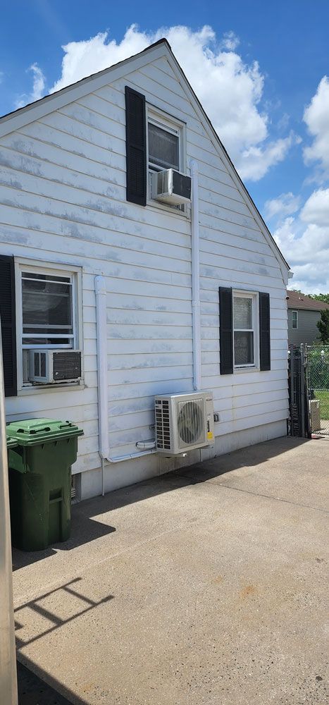 A white house with black shutters and a green trash can in front of it.