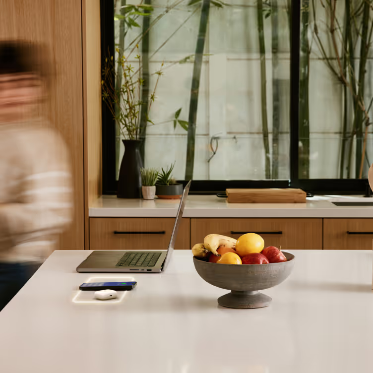 A kitchen counter with a laptop, fruit bowl, and charging station. A person is blurred in the background.