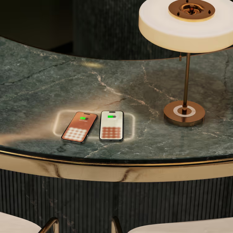 Two phones charging on a marble counter with a built-in wireless charger, next to a small lamp with a gold base and white shade.