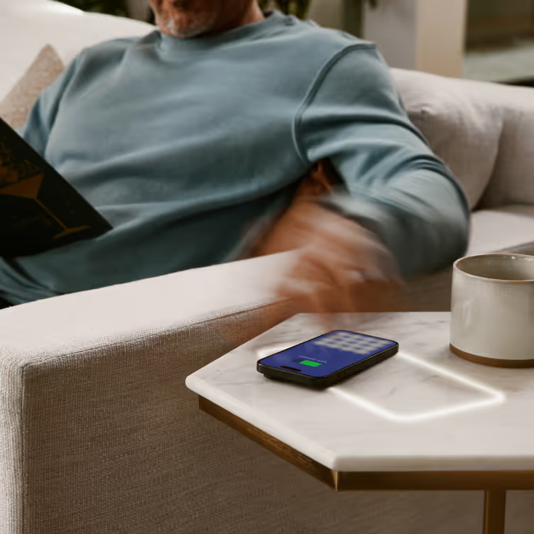 A man reads on a couch near a side table with a phone charging wirelessly. The table has a glowing outline.