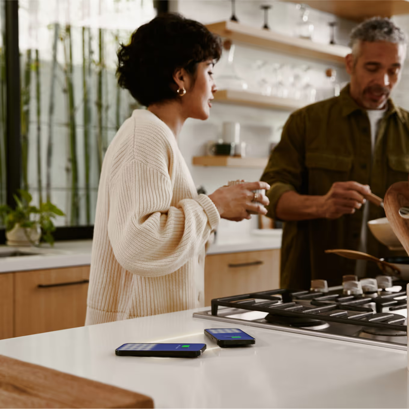 Woman and man in kitchen, cooking. Two smartphones on the counter. The woman is talking, and the man is stirring a pan.