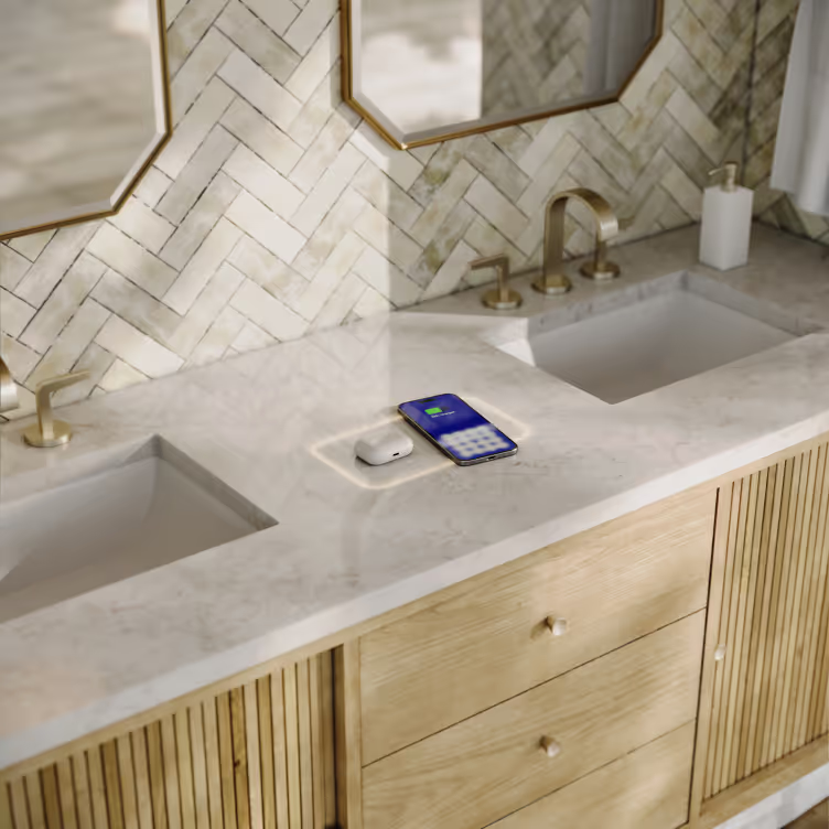 Bathroom vanity with two sinks, marble countertop, wooden cabinets, and gold fixtures; a phone and wireless earbuds are charging on a built-in wireless charging pad.