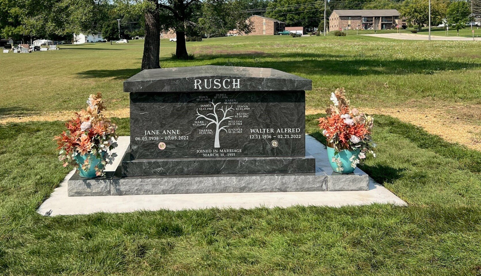 Granite Busch family tombstone with floral arrangements in a grassy cemetery.