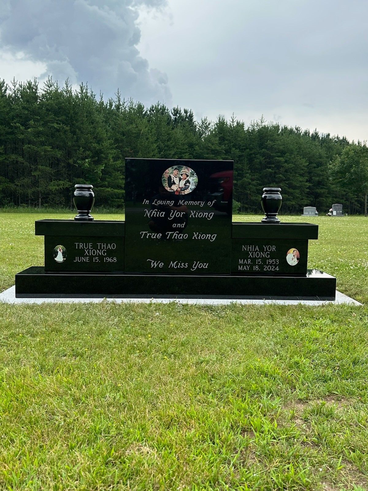 Black pet memorial with urns, engraved names, and photos of pets in a grassy field with trees.