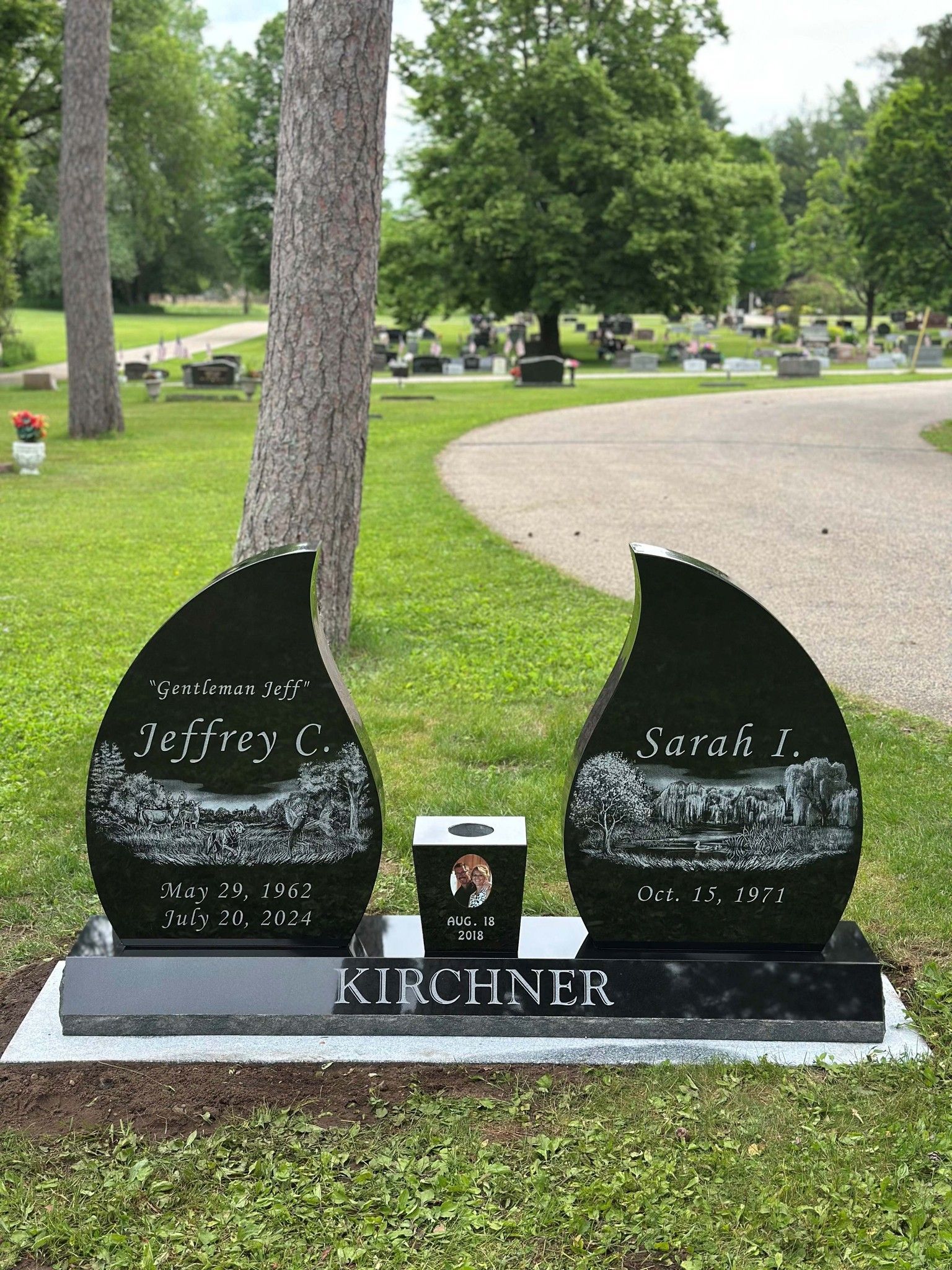 Black teardrop-shaped headstones in a cemetery, inscribed with names and dates, a photo urn between them.