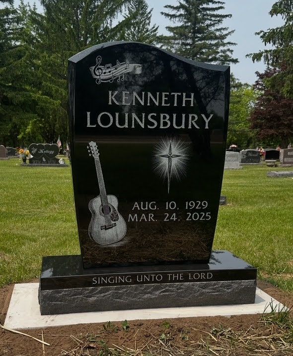 Black granite tombstone with guitar, cross, name, and dates, set in a cemetery.