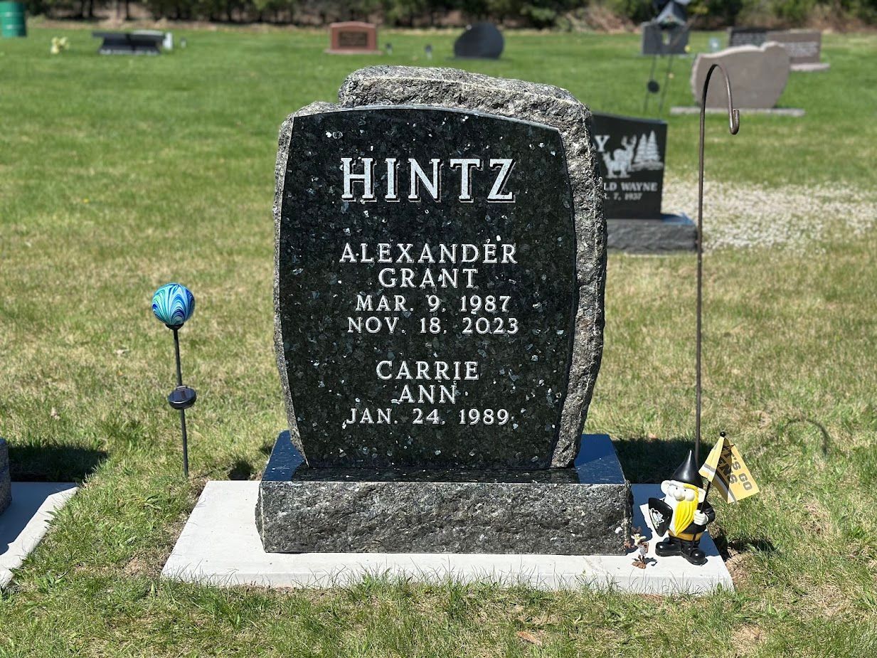 Tombstone in a cemetery.  Reads: HINTZ, Alexander Grant (1987-2023) and Carrie Ann (1989- ).