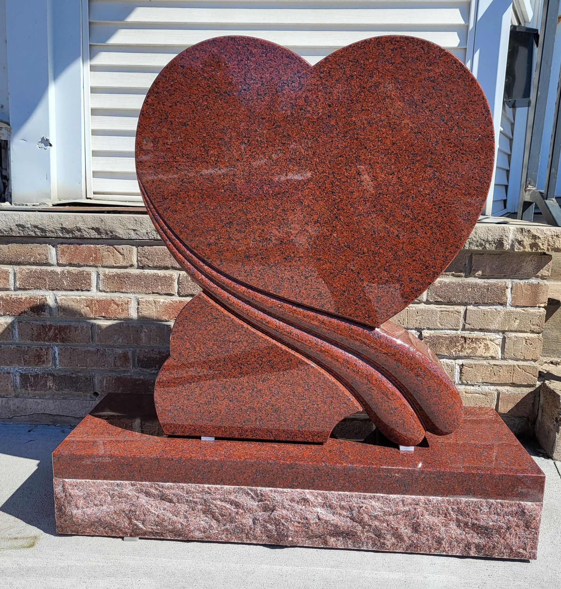 Red heart-shaped granite headstone on a matching base in front of a building.