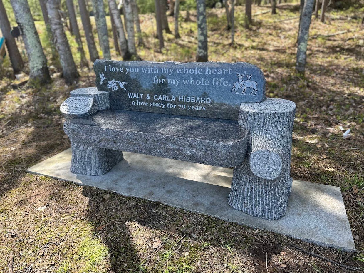 Granite bench inscribed with a memorial, in a wooded area.
