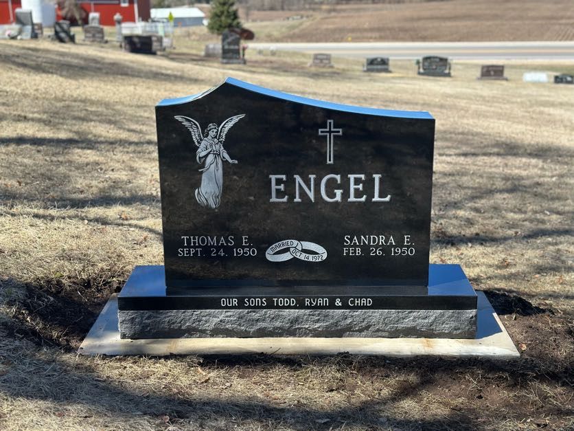 Black Engel headstone with angel, cross, names, dates, and wedding rings in a cemetery.