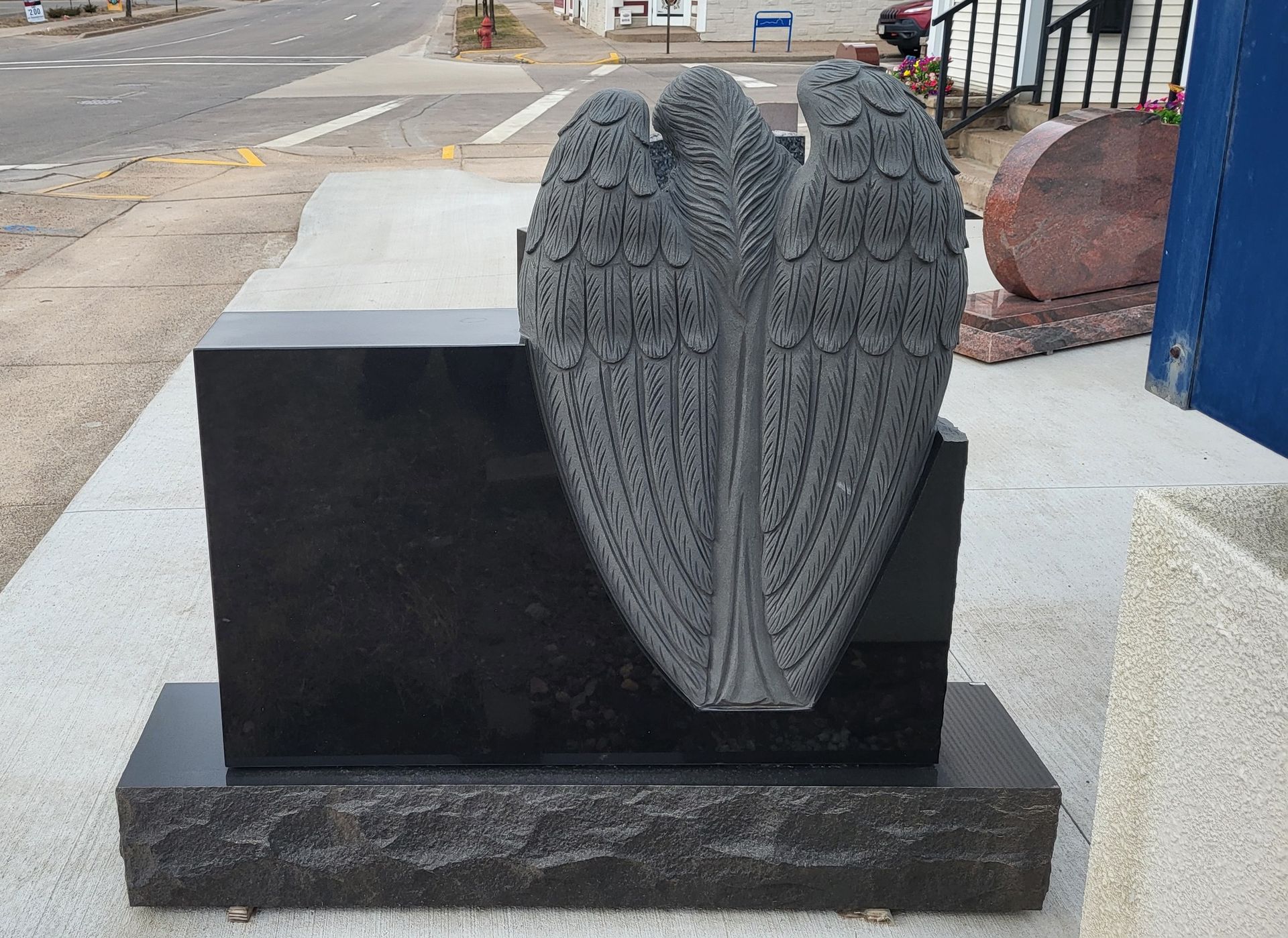 Black granite headstone with angel wing carving, on a sidewalk.
