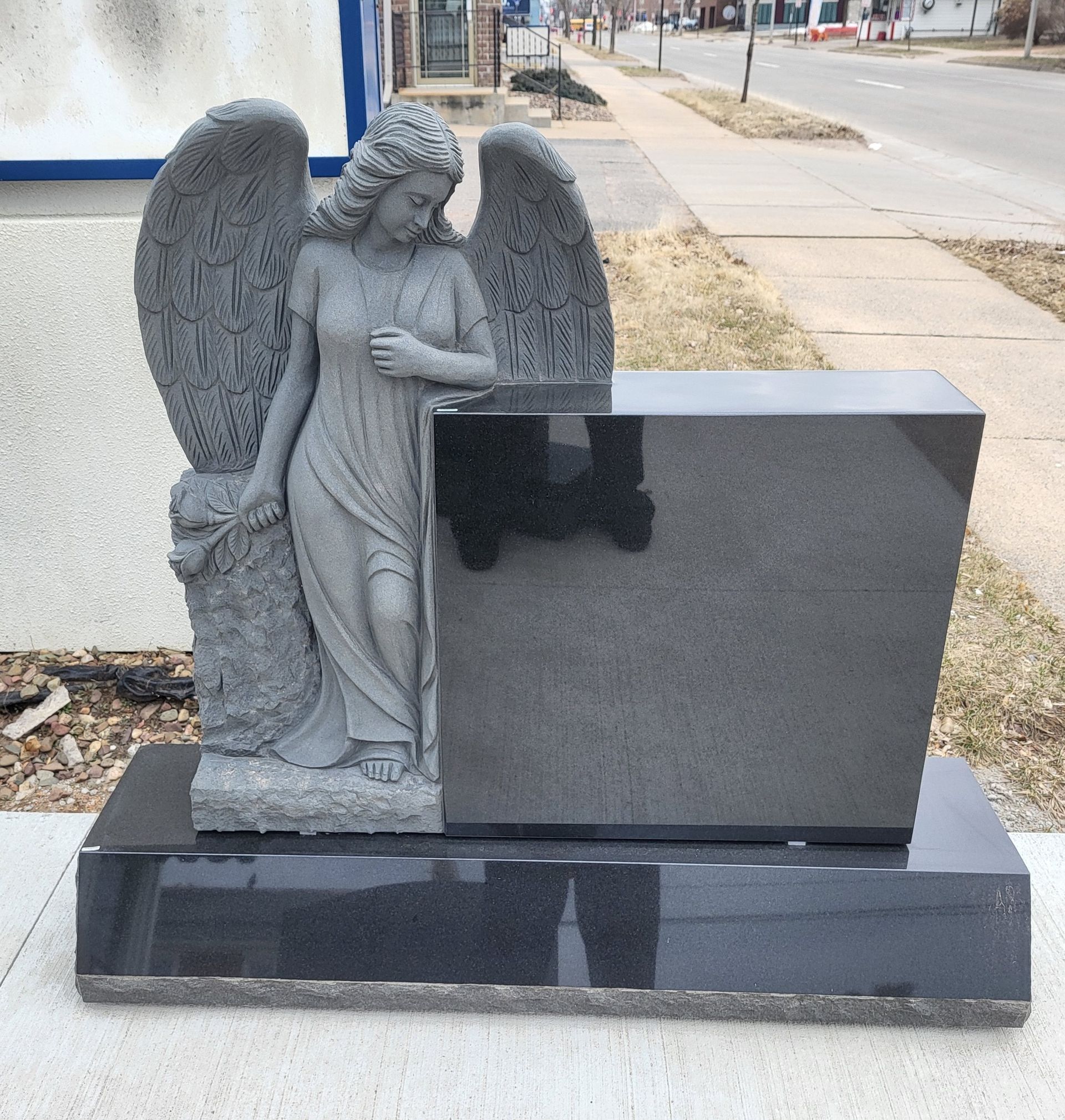 Gray angel statue beside a black granite tombstone, set on a sidewalk.
