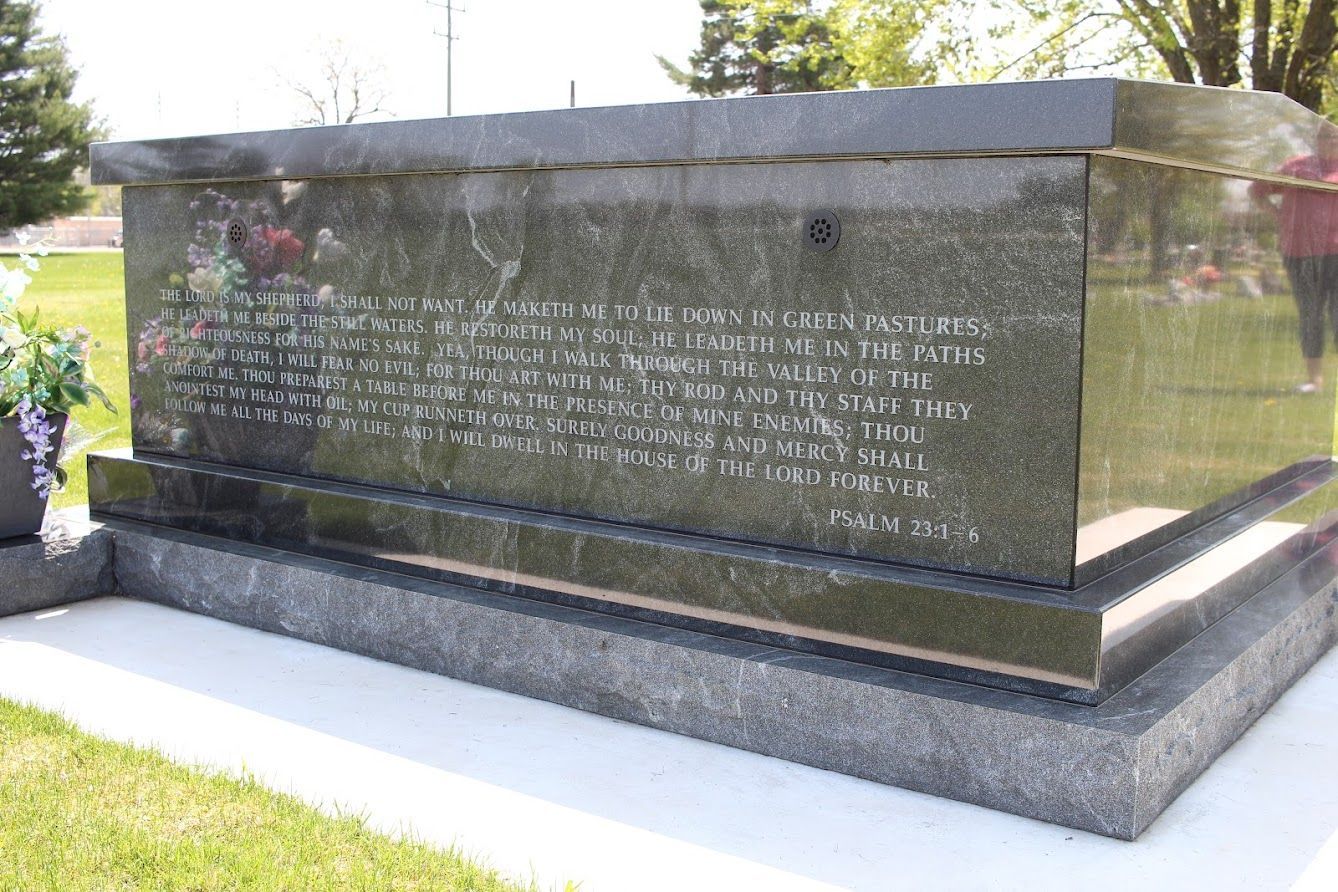 A large, dark granite tomb with an inscription in a cemetery; a person stands nearby.