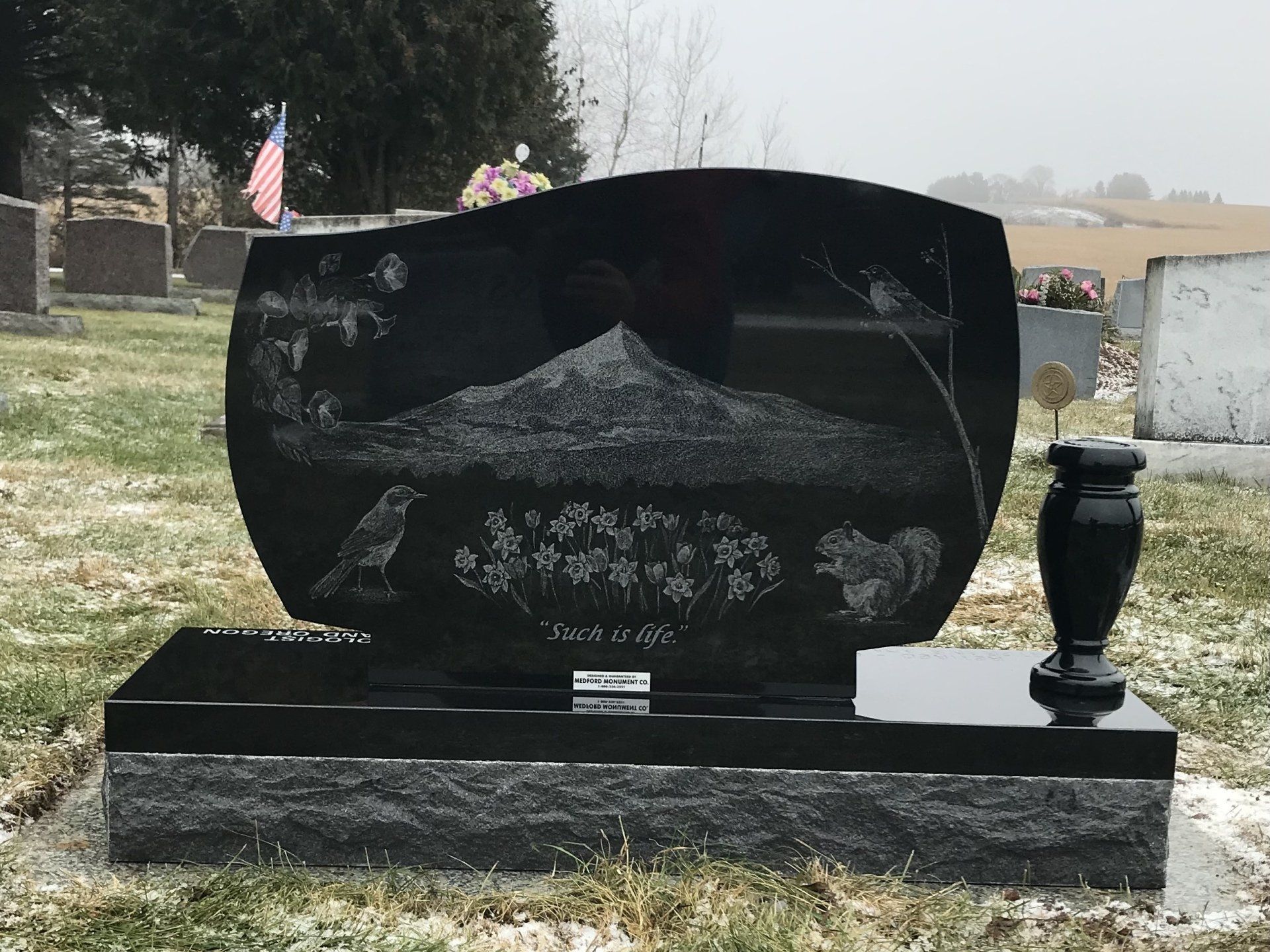 Black gravestone with etched mountain scene, bird, and vase in a snowy cemetery.