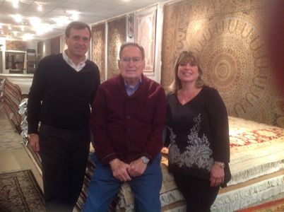 Three people pose among stacks of rugs in a rug store. Man in the center sits with arms crossed, flanked by a man and woman.