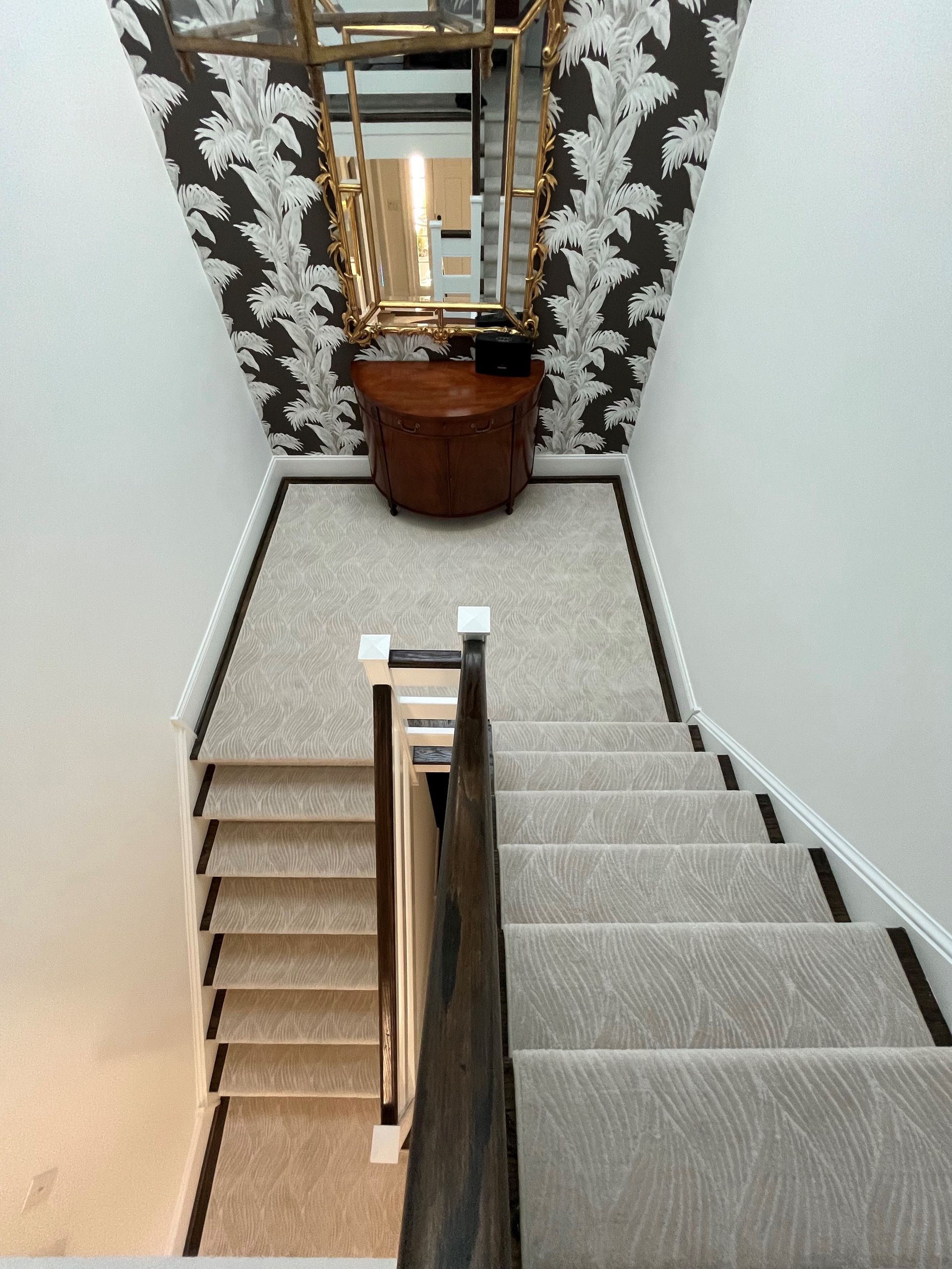 A staircase with gray carpet and a black border leads to a landing with wallpaper, a mirror, and a wooden table.