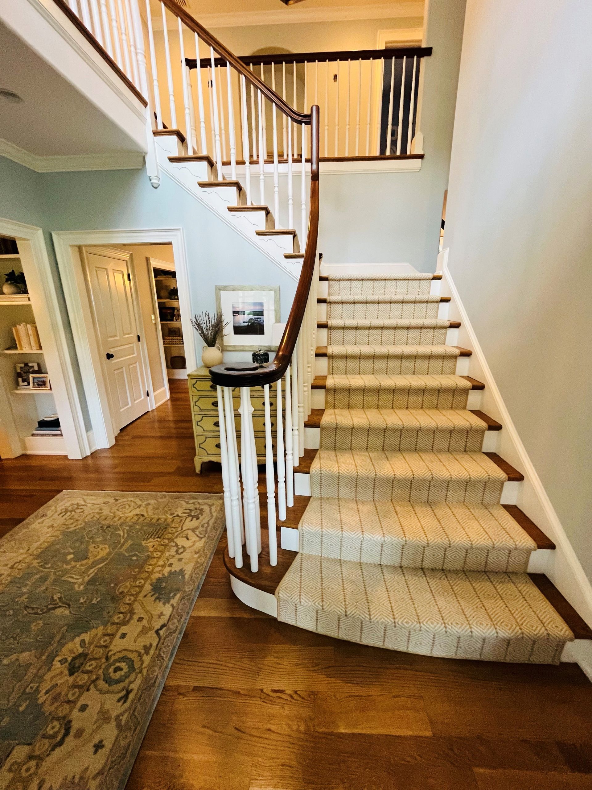 A two-story foyer with a staircase. Stair treads are carpeted; walls are pale blue. A runner and wooden floors are below.