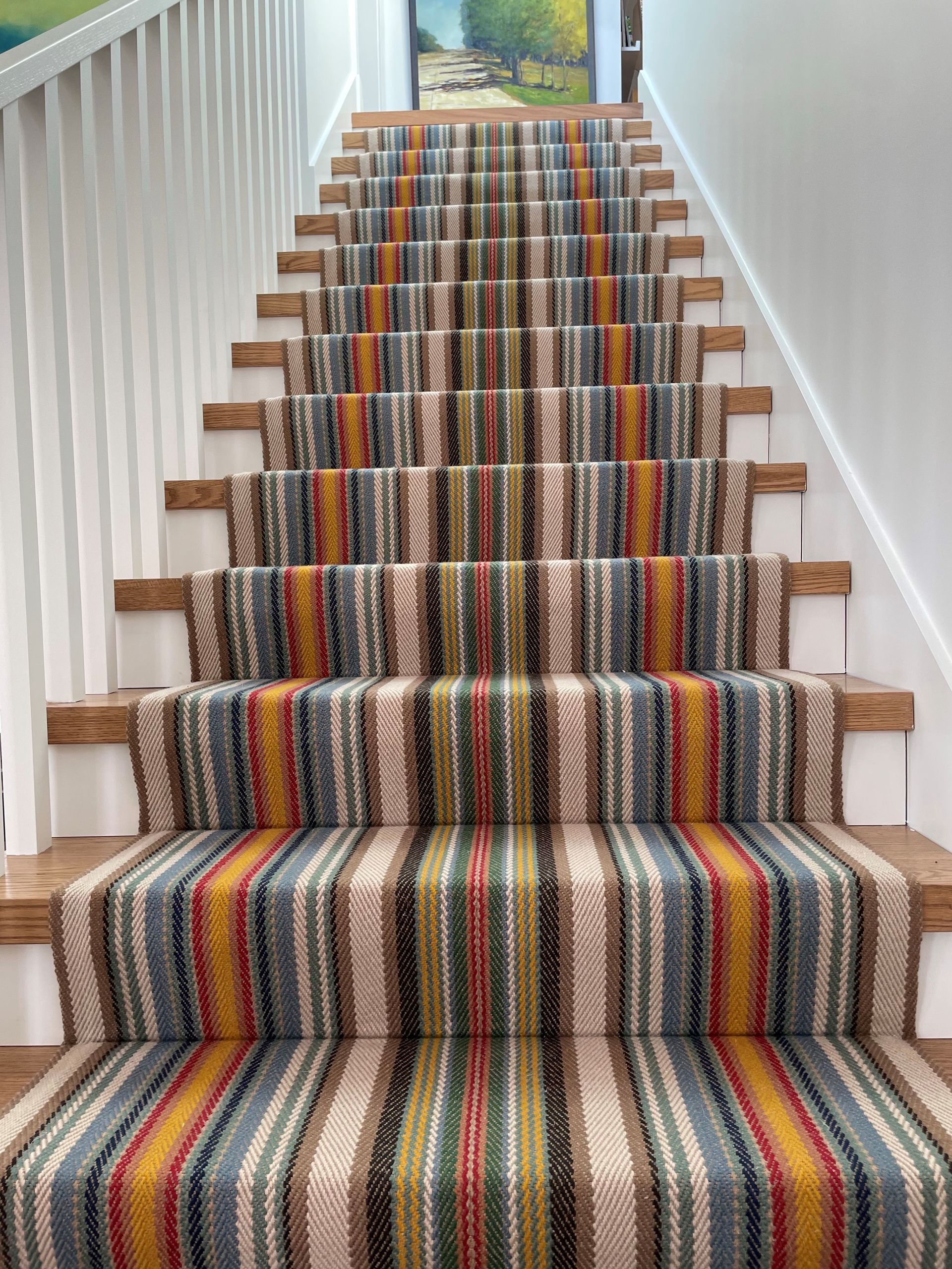 A set of wooden stairs with a colorful striped carpet runner; the walls are white.