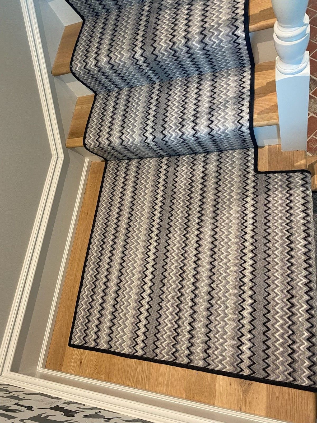Staircase with patterned carpet runner in shades of gray, black, and white, bordered by wooden stairs and a white column.