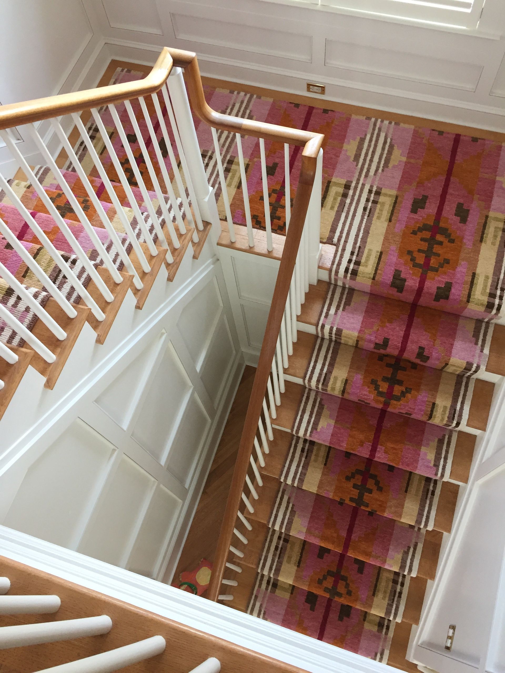 Staircase with a pink and brown patterned runner, white paneling, and wooden handrails. Top-down view.