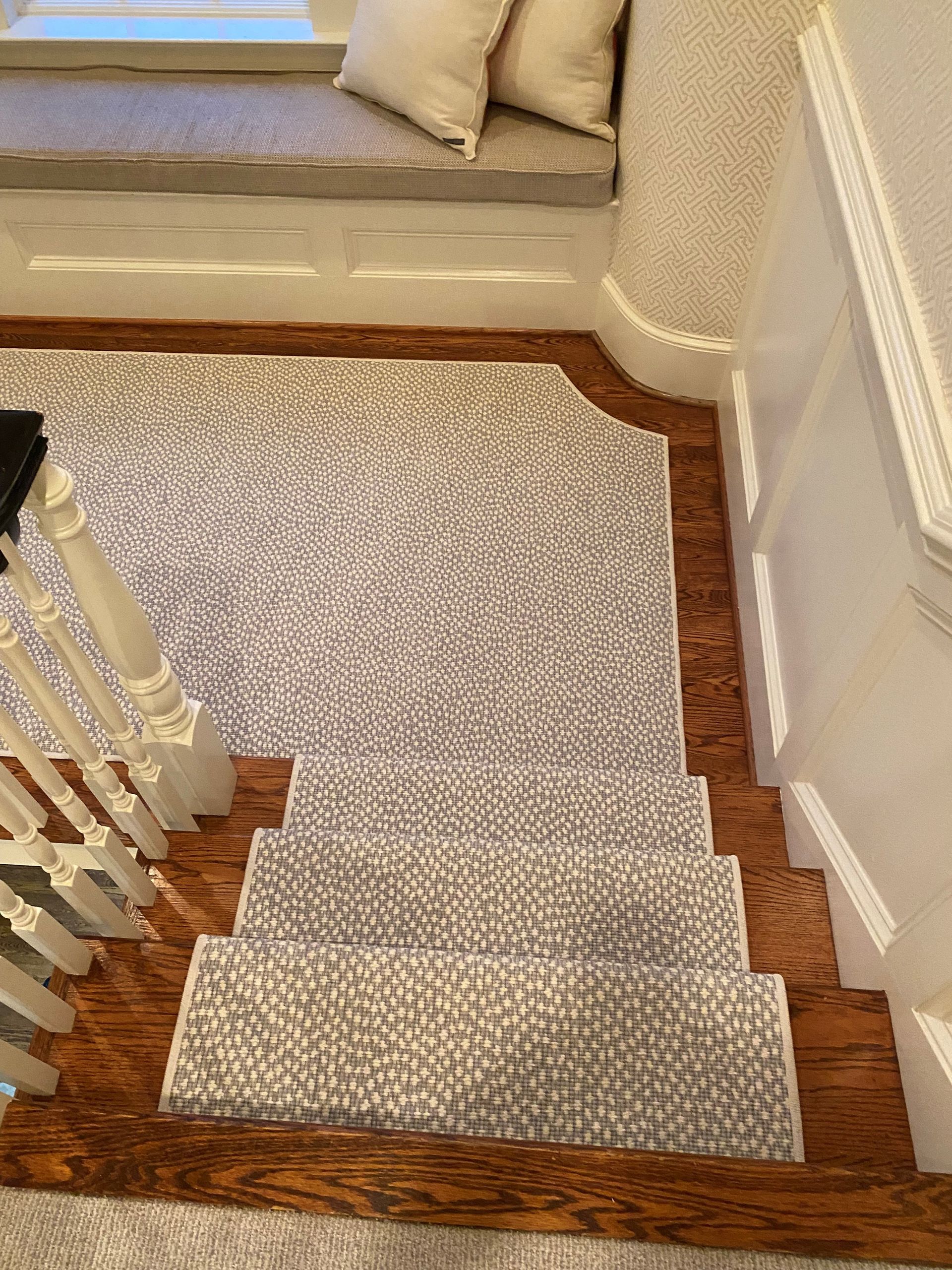 Overhead view of a staircase with a gray patterned carpet runner on wood steps. A window seat with pillows is in the background.