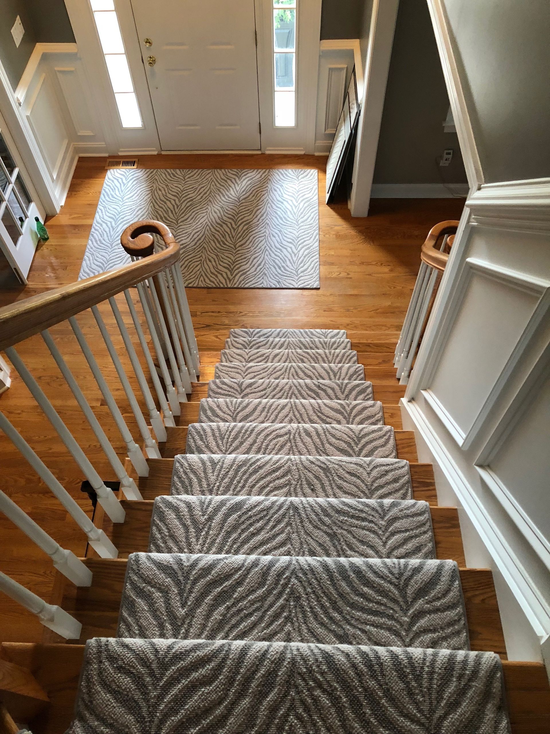 Staircase with patterned carpet runner leads to a hardwood foyer with a neutral rug and front door.  A railing and wall paneling are visible.