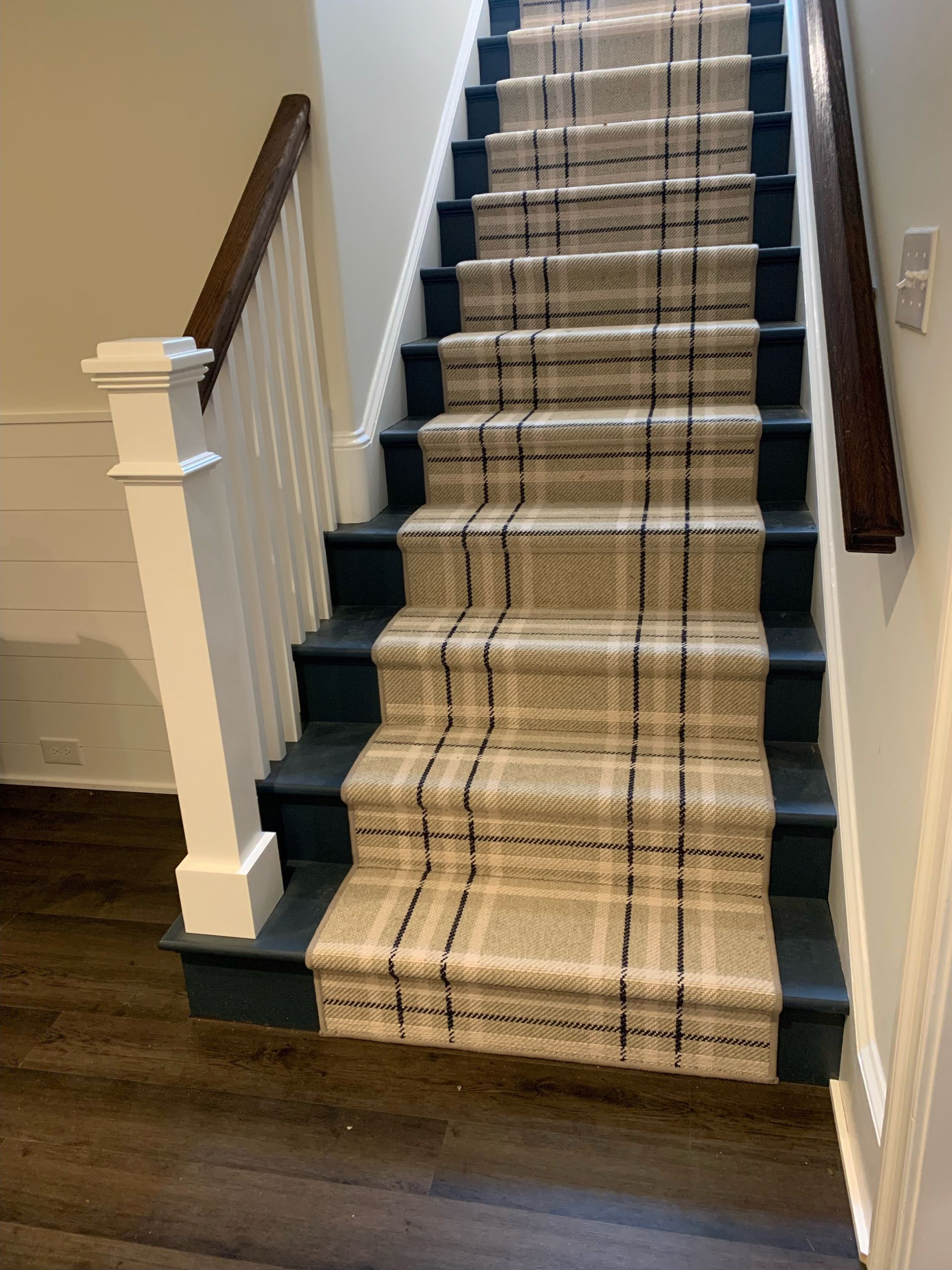 Staircase with tan and black plaid carpet, dark blue risers, white balusters, and a dark wood handrail.