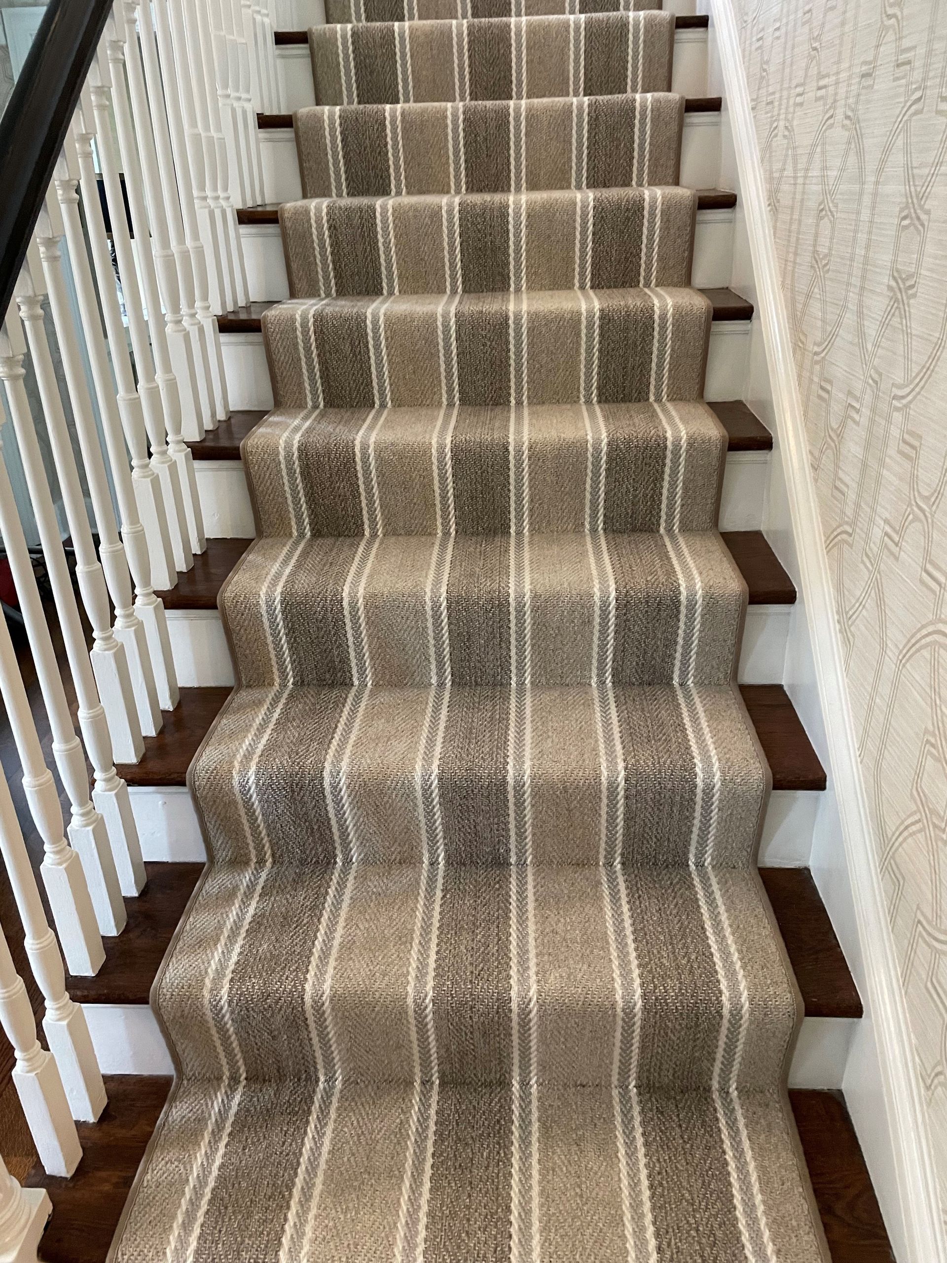 Staircase with a carpet runner in shades of brown and white. White railing on the left and textured wallpaper on the right.