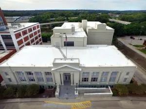 White building with columns, steps, and flagpole; surrounded by other buildings and greenery.