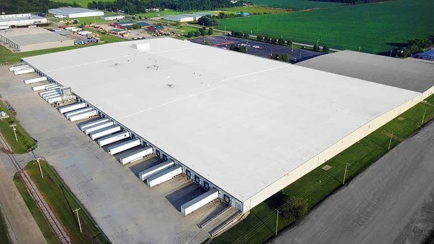 Aerial view of a large warehouse with loading docks, surrounded by pavement and green fields.