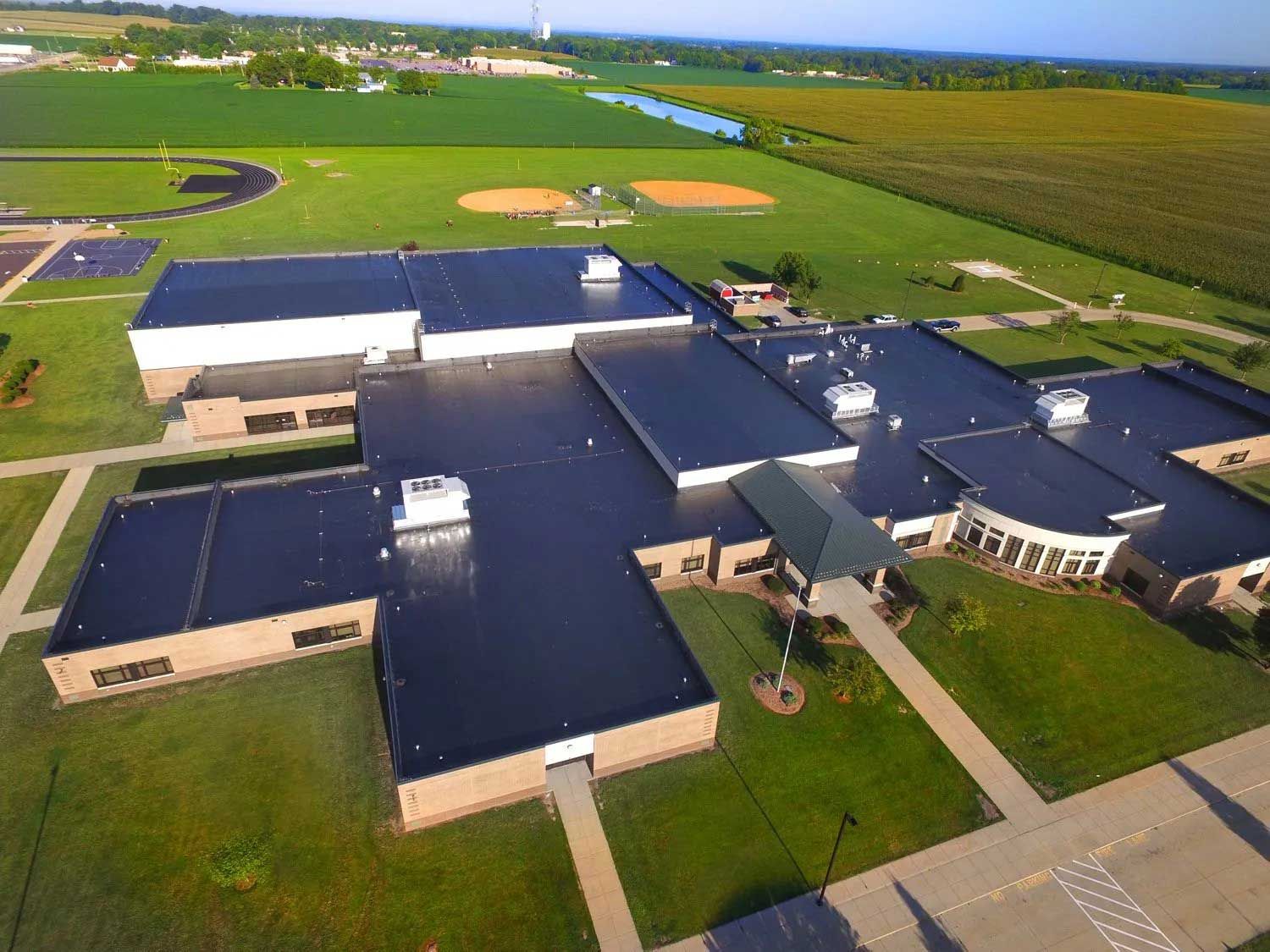 Aerial view of a school building with a dark roof and surrounding green fields, including sports fields.