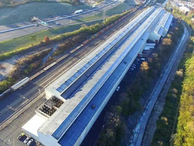 Aerial view of a long, industrial building beside train tracks and a highway, surrounded by green vegetation.