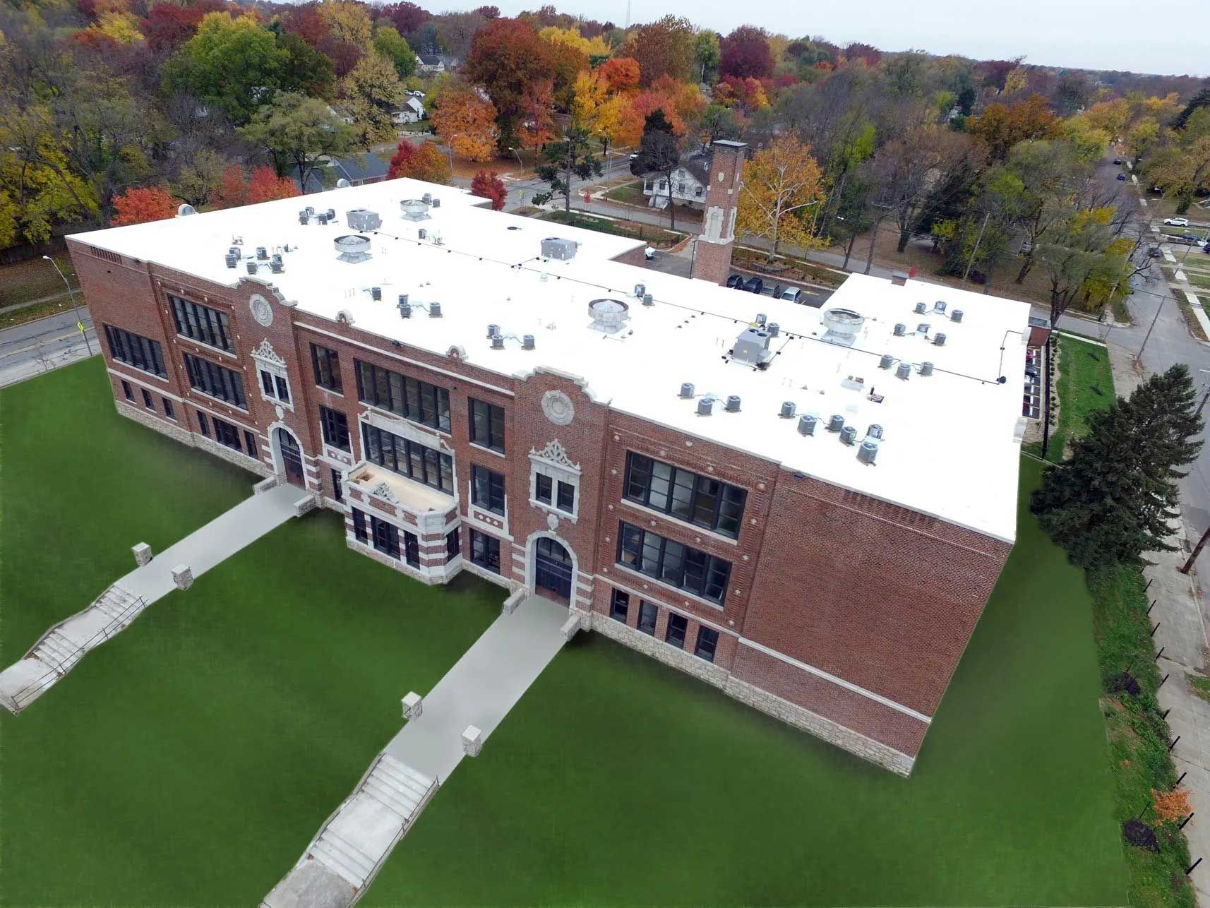 Aerial view of a brick school building with a white roof and green lawn, surrounded by colorful autumn trees.