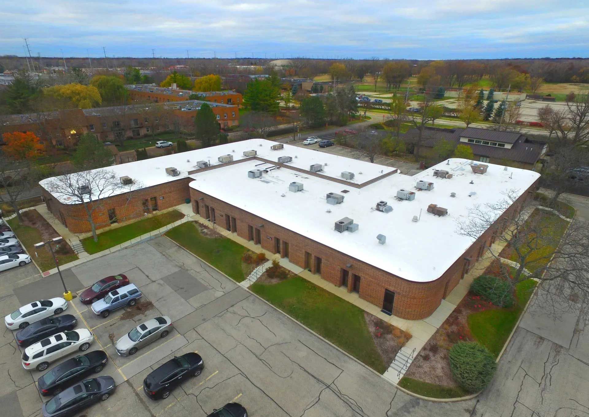 Aerial view of a one-story brick building with a white roof, surrounded by a parking lot and trees.
