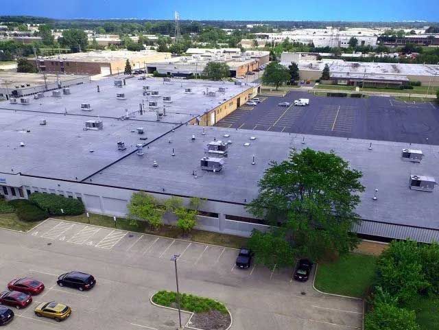 Aerial view of a large industrial complex with flat roofs, parking lot, and surrounding greenery. Cars are parked.