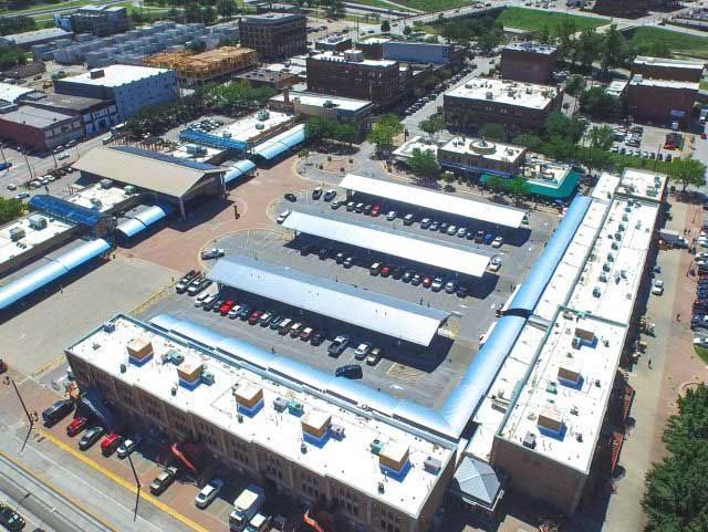 Aerial view of a city block with buildings, parking areas, and cars. Structures have flat roofs and parking canopies.