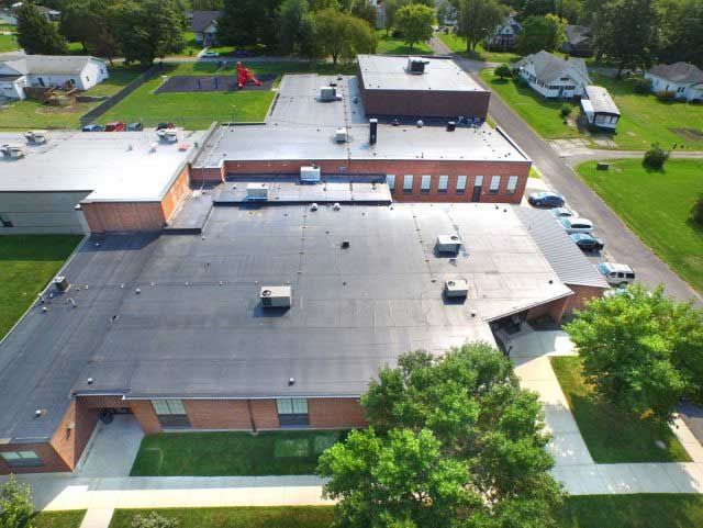 Aerial view of a school building with a flat roof, brick exterior, and surrounding green lawn and trees.