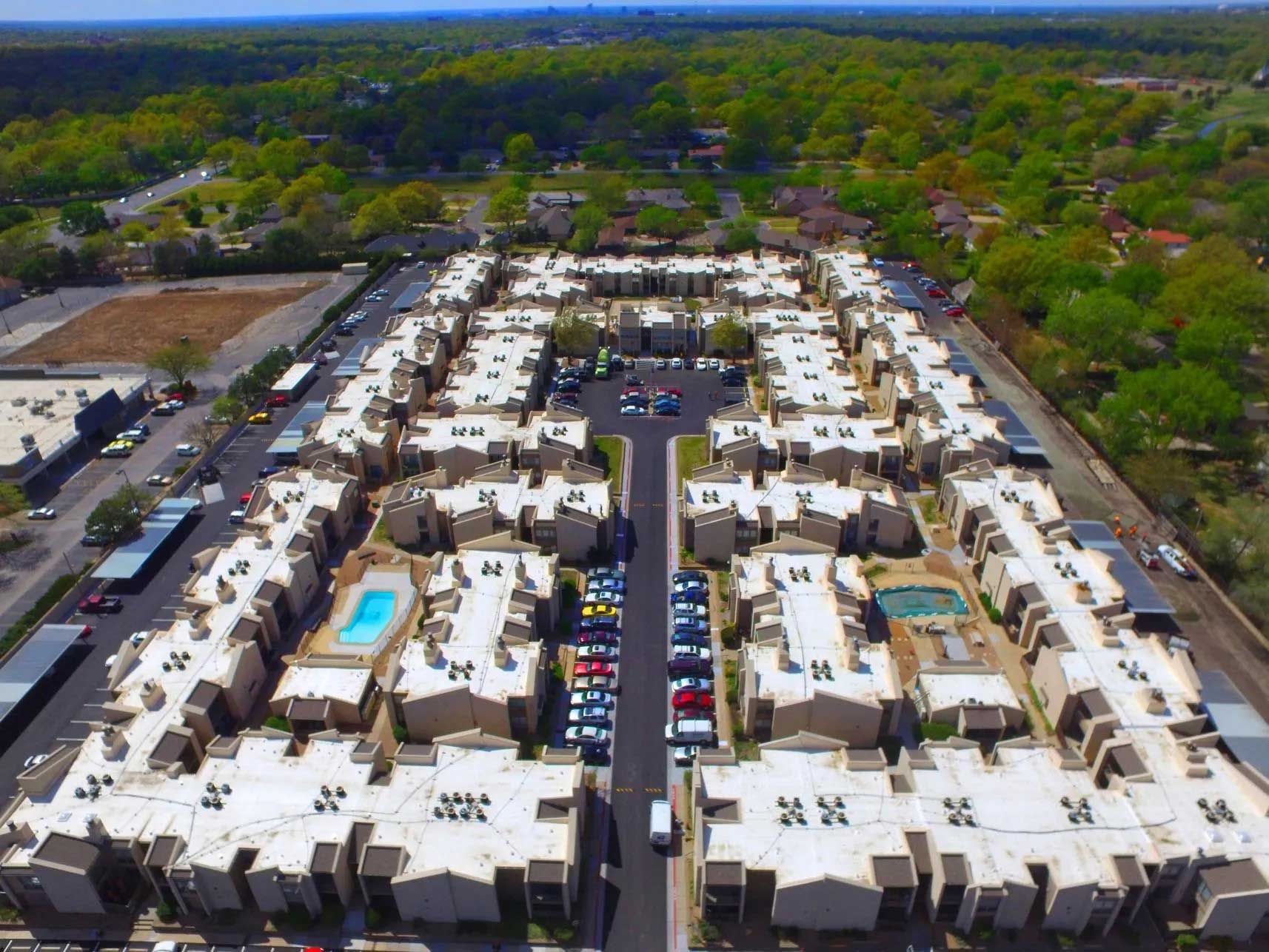 Aerial view of a large apartment complex with parking, pools, and surrounding trees.