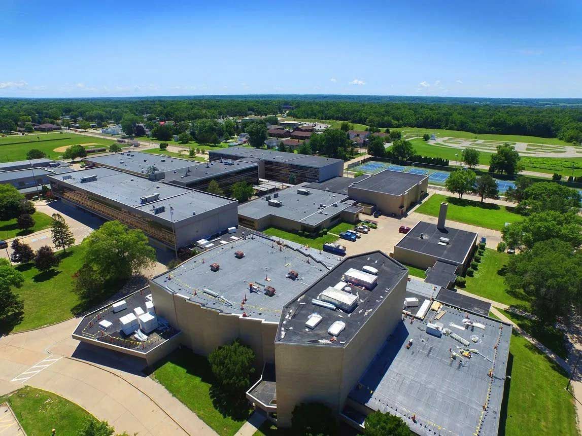 Aerial view of a school campus with gray buildings, green grass, and trees under a blue sky.