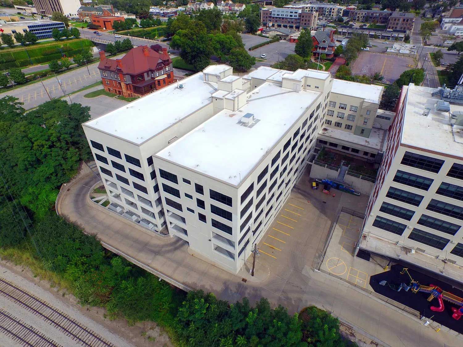 Aerial view of a white multi-story industrial building with many windows and flat roofs, located beside greenery and a road.