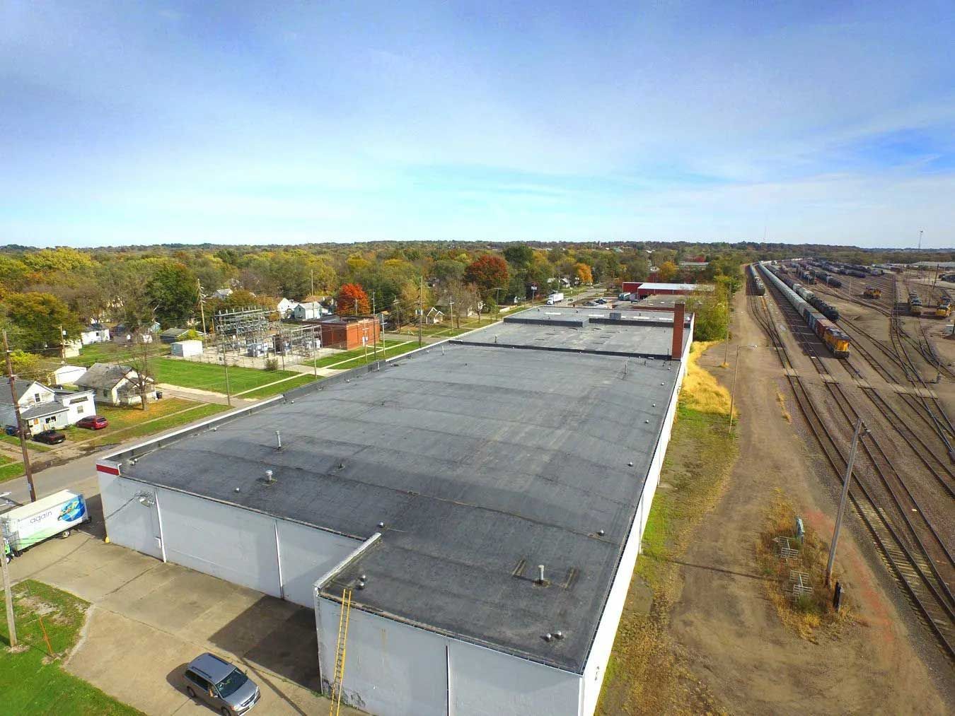 Overhead view of a large industrial building, with a flat roof, next to train tracks under a blue sky.