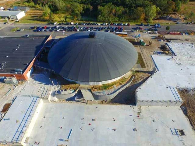 Aerial view of a large, dark dome-shaped building under construction with surrounding concrete and parking lot.