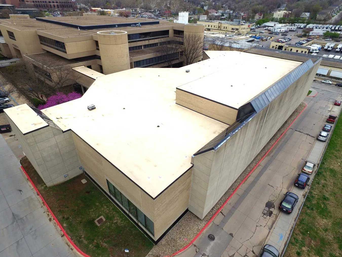 View of a large, beige institutional building with a flat roof. A road and parked cars are along the side.
