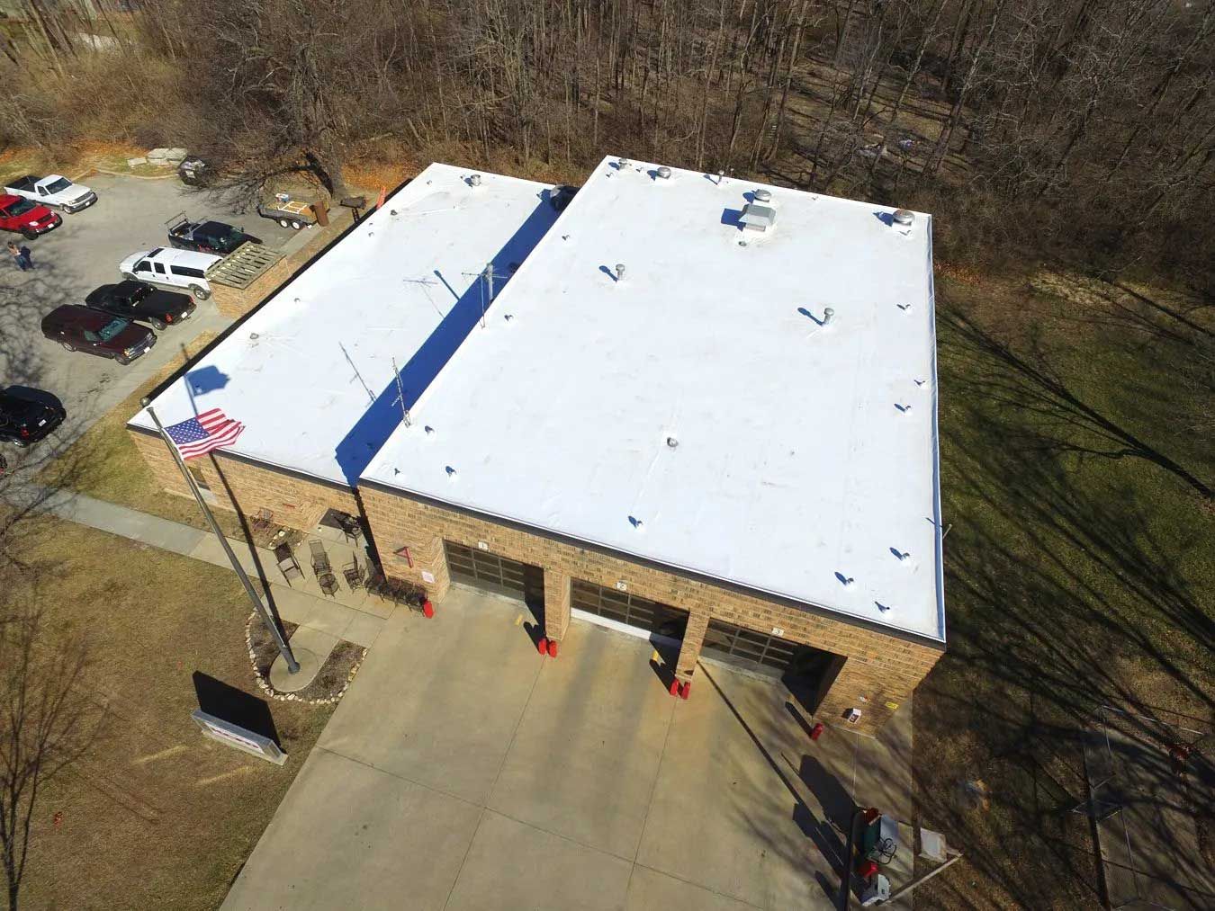 Aerial view of a fire station with a white roof, three garage doors, and American flags in front. Cars parked nearby.