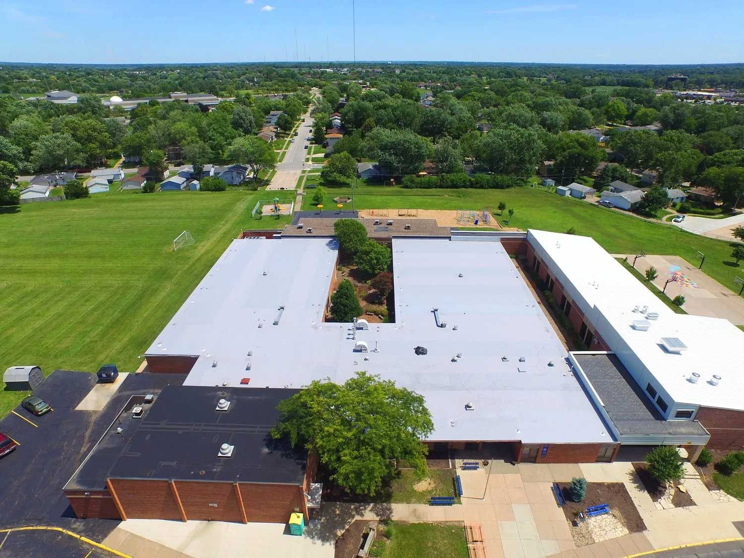 Aerial view of a school building with a gray roof and a green playing field, surrounded by trees and a neighborhood.