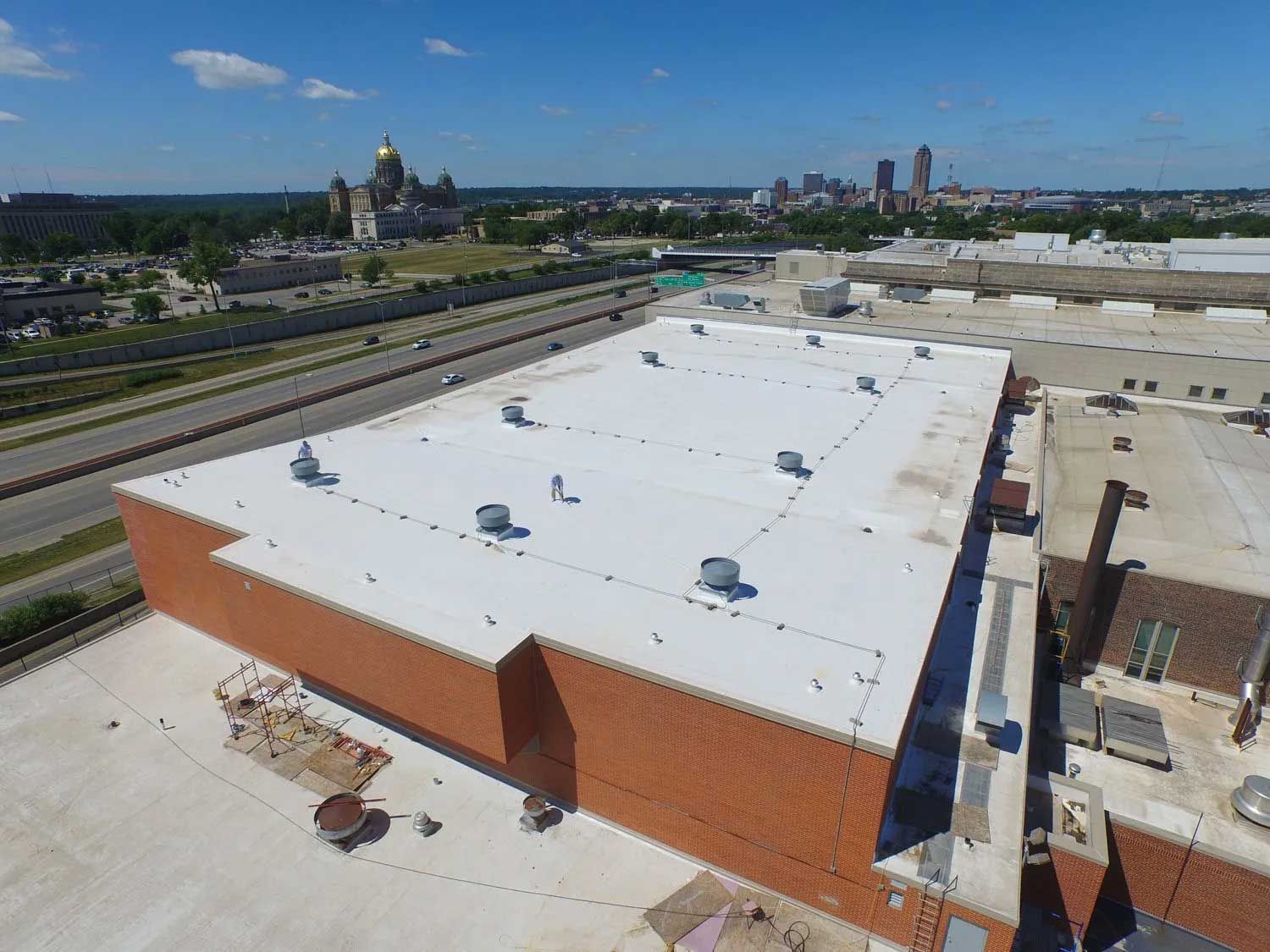 Aerial view of a large brick building with a white roof, city skyline in the background, blue sky.