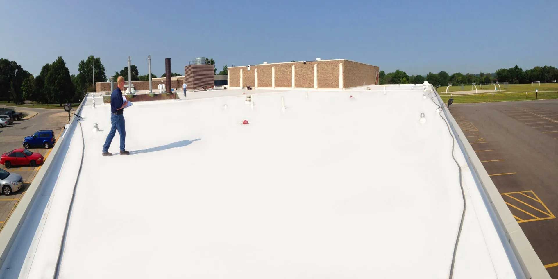 Man on a white roof inspecting the surface. Building and parking lot visible under blue sky.