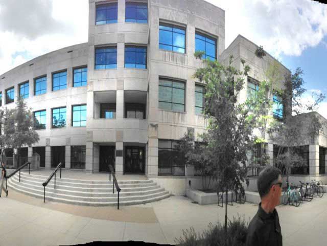 Stone building with blue-tinted windows, steps, and trees. Person walks by in the foreground.