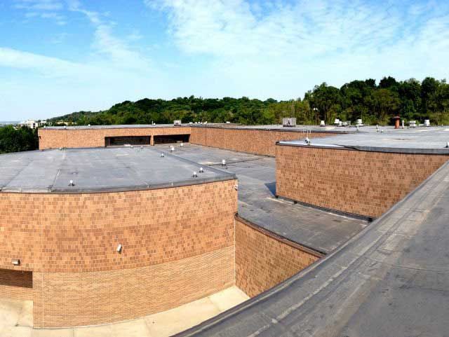 Brown brick building with flat black roof and trees in the background under a blue sky.