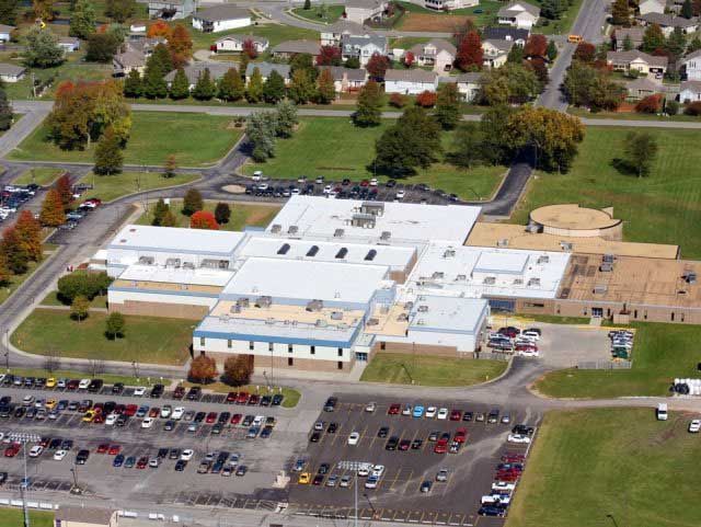 Aerial view of a school building with a flat roof, surrounded by parking lots, trees, and residential neighborhood.